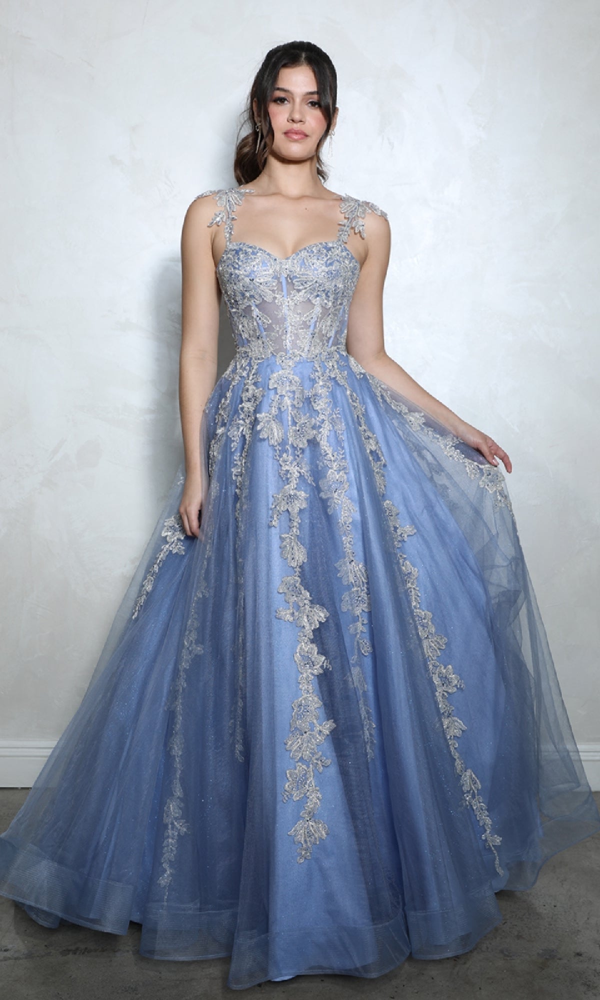 A woman in the Long Blue Sweetheart Prom Gown: Eureka EK115, featuring silver embroidered floral details, stands against a light-colored textured wall.